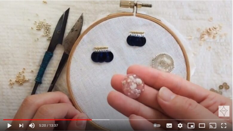 刺繍枠の前で、作家さんが作ったカスミソウが閉じ込められている樹脂パーツを手に乗せて紹介している写真