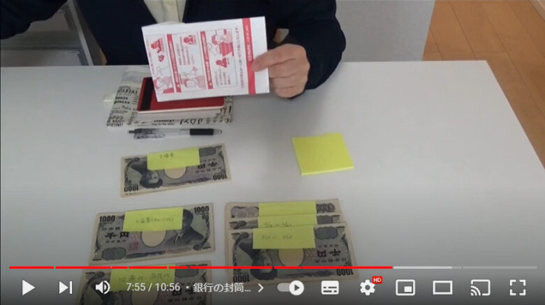 テーブルには付箋紙で挟まれた千円札の束がいくつか並んでいます。銀行においてある封筒を片手でつまんでいます。