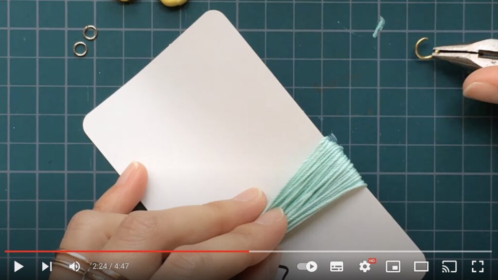 てのひらサイズの白い厚紙に緑色の刺繍糸を何重にも巻いているようすを写した写真。タッセルを作るために、右手にペンチを持って丸カンを挟んでいます。