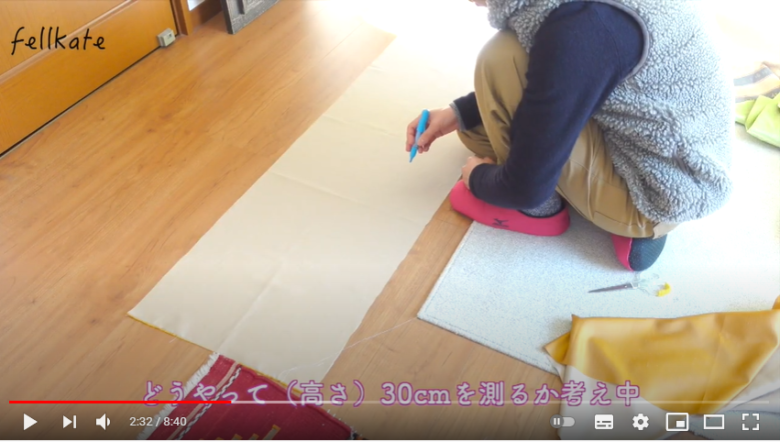 チャコペンを手に床に敷いた布地の前に座り、布地をどう必要な大きさに切る方法を考えている様子
