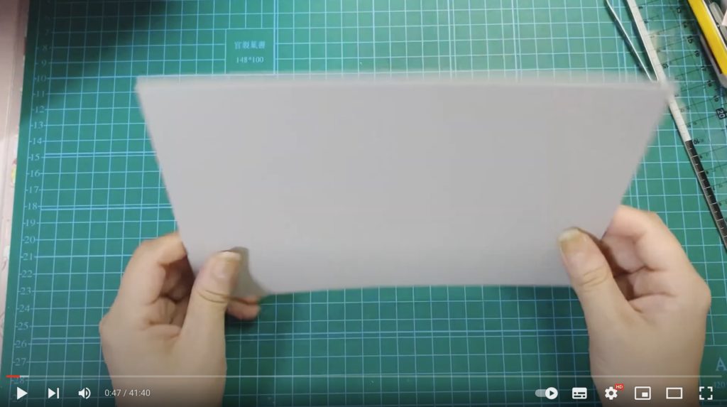 シェーカーづくりに使用している白い厚紙を両手に持って紹介している写真。緑色のボードの上で作業をしています。