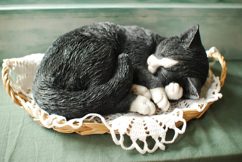 白い布が敷かれたカゴの上に黒と白の模様の猫の置物が寝転がっている様子を写した写真。