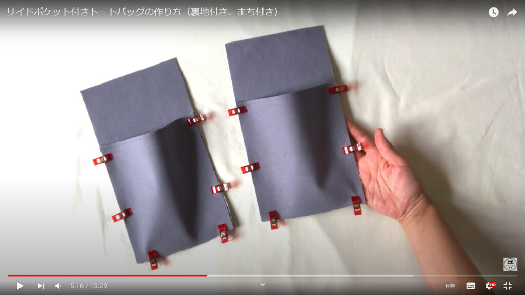 サイドポエット部分と本体の作る作業を解説している動画で、机に置かれたクリップで留めた2つのサイドポケットを手に取った画像。