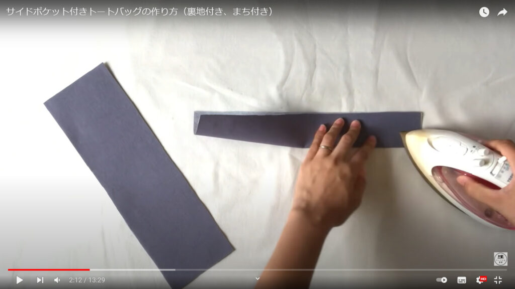 持ち手の部分を作る作業を解説している動画で、持ち手の部分の生地を折ってアイロンを当てている画像。