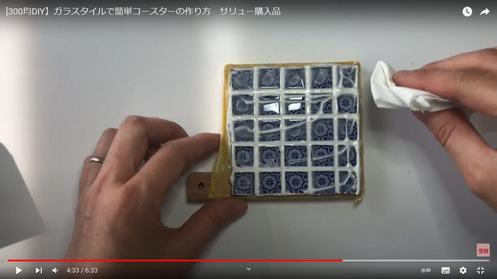 仕上げの拭きあげ作業を説明している動画で、机に置かれた目地材を塗ったコースターを手で押さえ、もう片方の手に小さく畳んだティッシュを持った画像。