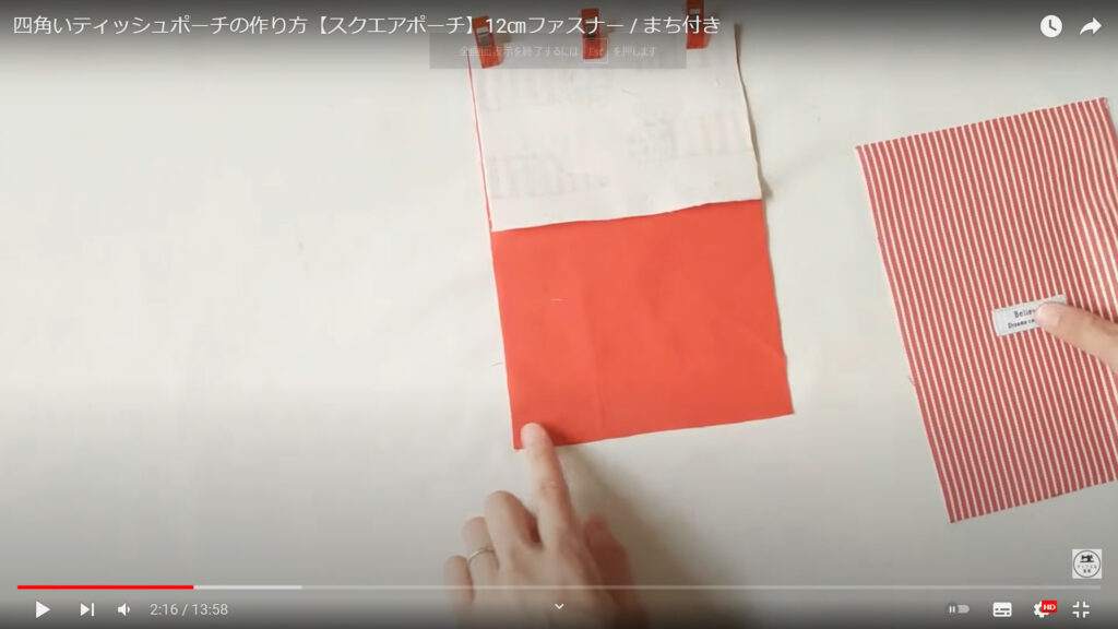 ポーチのポケットティシュの部分を作る作業を解説している動画で、表地とポケットティシュの裏地を合わせたものを指さし、もう1枚の表地を手にしている画像。