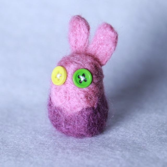 ピンクパープルの一頭身のウサギの羊毛マスコットの画像です。目には緑と黄色のボタンが使われています。