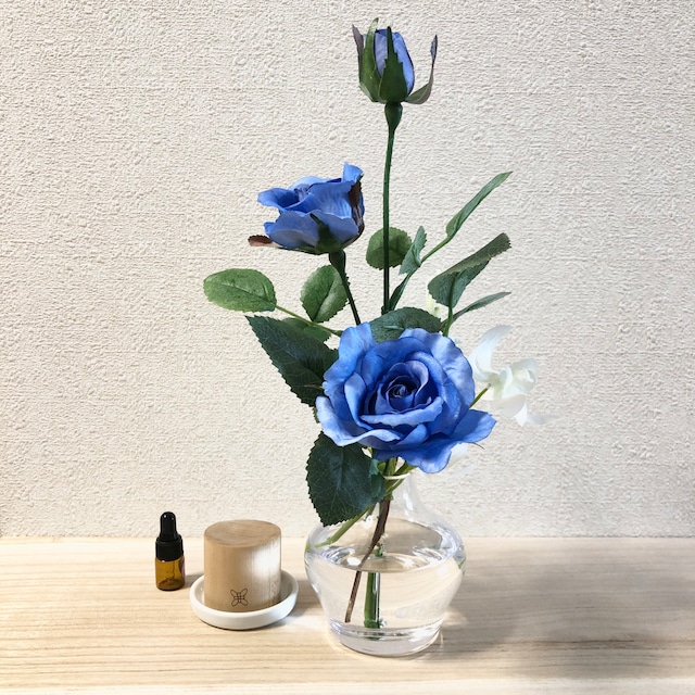 青いバラとデラニウムなどが透明の花瓶に活けられており、エッセンシャルオイルとアロマウッドと一緒に写っている写真。