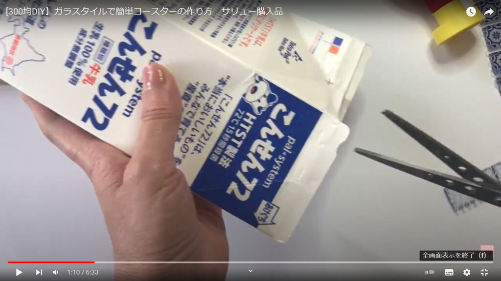 ガラスタイルのコースターで使用する牛乳パックのへらの作り方を説明している動画で、牛乳パックを手に持ち、ハサミでカットする様子を表示した画像。