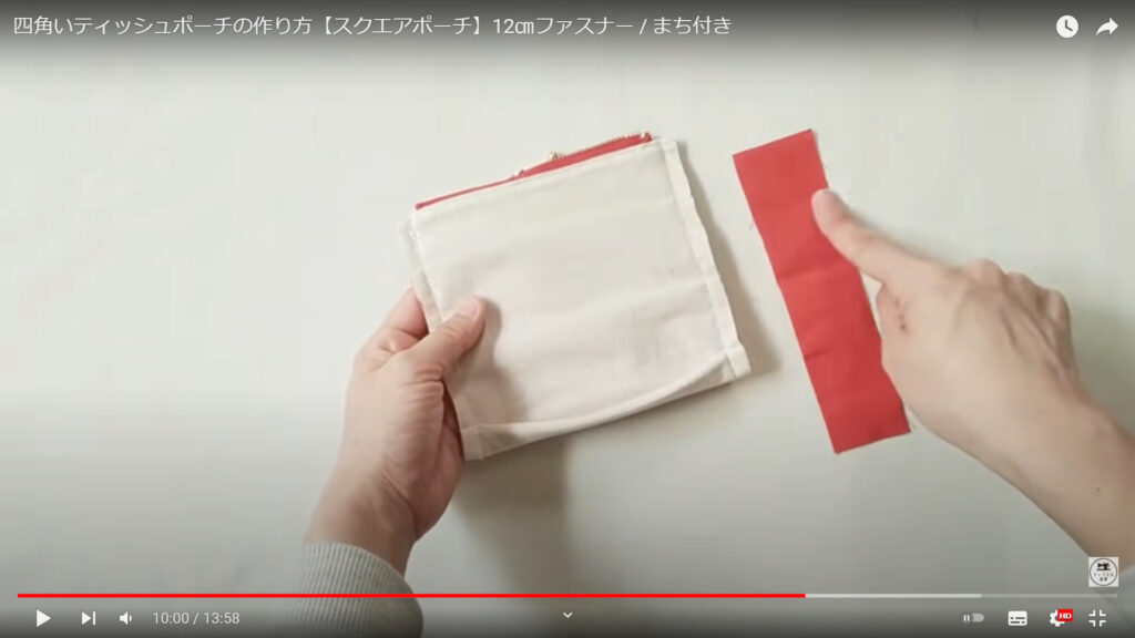 ポーチの端の処理する作業を解説している動画で、マチを作ったポーチ本体を片手に、端に縫い付ける小さめの赤い生地を指さした画像。