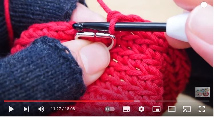 指先が出るタイプの黒い手袋をした手で赤色の編地にシルバーのDカンを編みつけるところの画像