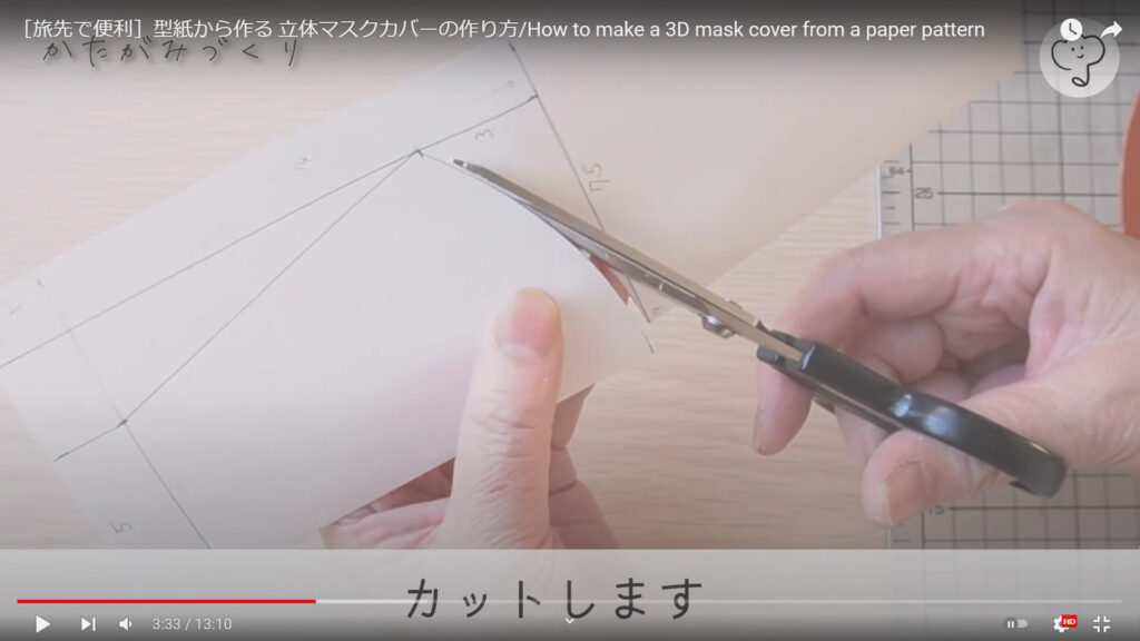 マスクカバーの型紙を作る作業を説明している動画の途中で、紙に書かれた型紙をハサミで切り取っている様子と共に、「カットします」という文が表示された画像。