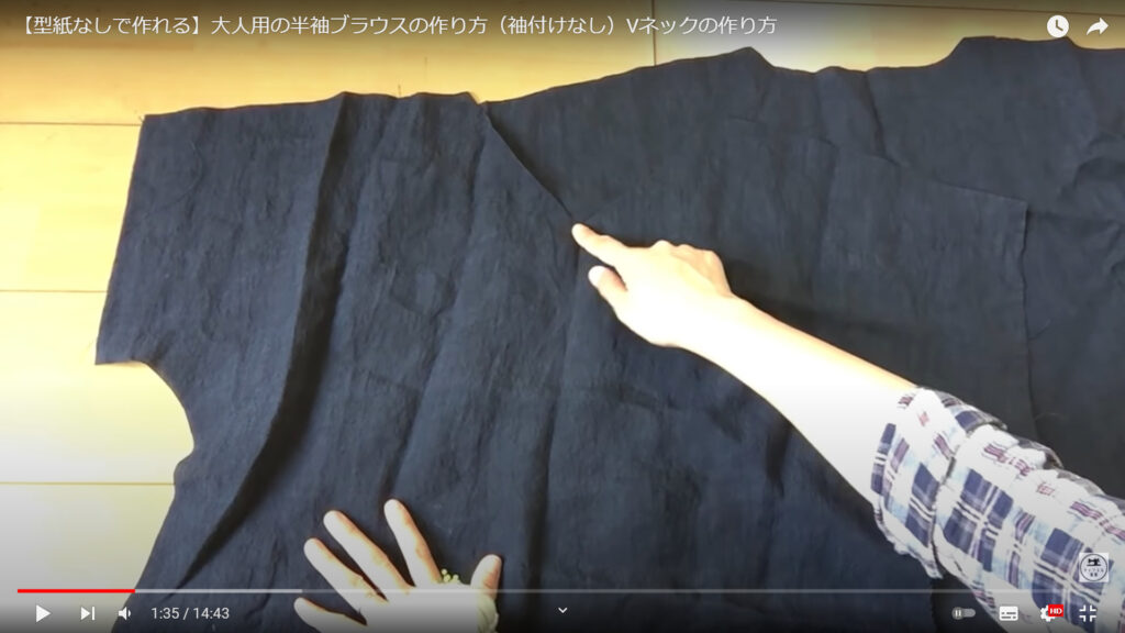 ブラウスの布を裁断する作業を解説している動画で、机の上にある裁断した前身ごろのVネックの部分を指さした画像。