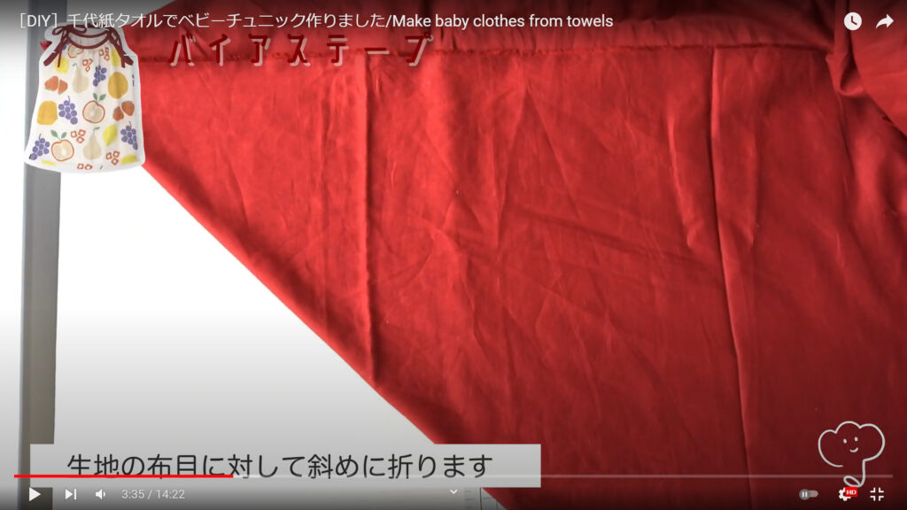 赤い生地からバイアステープを作る作業を説明した動画の途中で、「生地の布目に対して斜めに折ります」という文字が表示されている画像。
