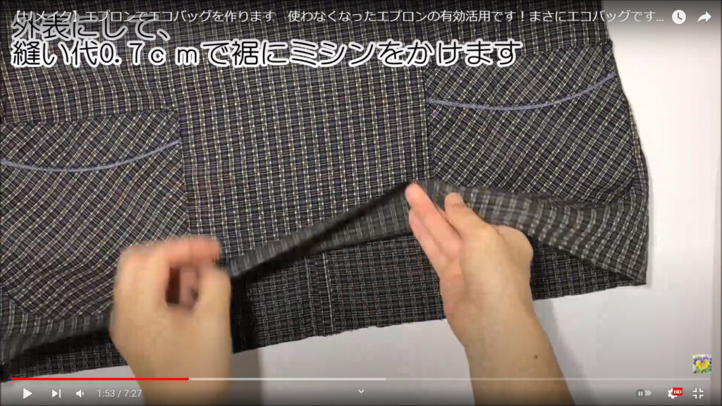 マチなしエコバッグの底の部分を縫い合わせる作業を解説している動画で、裾を手でめくっている様子と共に、「外表にして、縫い代0.7㎝で裾にミシンをかけます」という文が表示された画像。