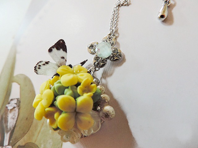たくさんの黄色い菜の花の上に紋白蝶がとまっている様子をデザインしたネックレスが写っている写真。