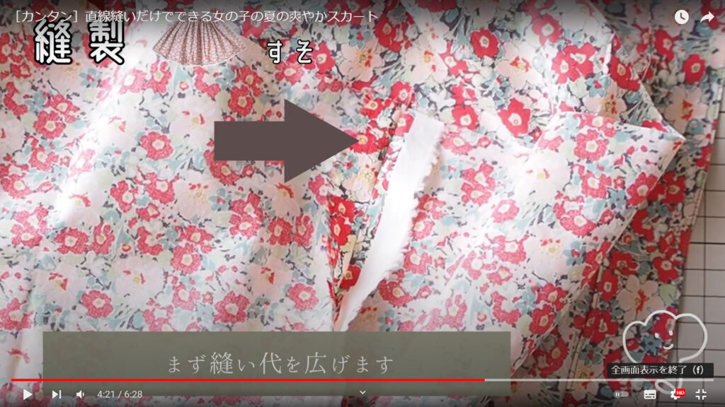 スカートの裾の部分を縫って仕上げる作業を解説している動画で、サイドの縫い代を広げたところを矢印で示し、「まず縫い代を広げます」という文が表示された画像。