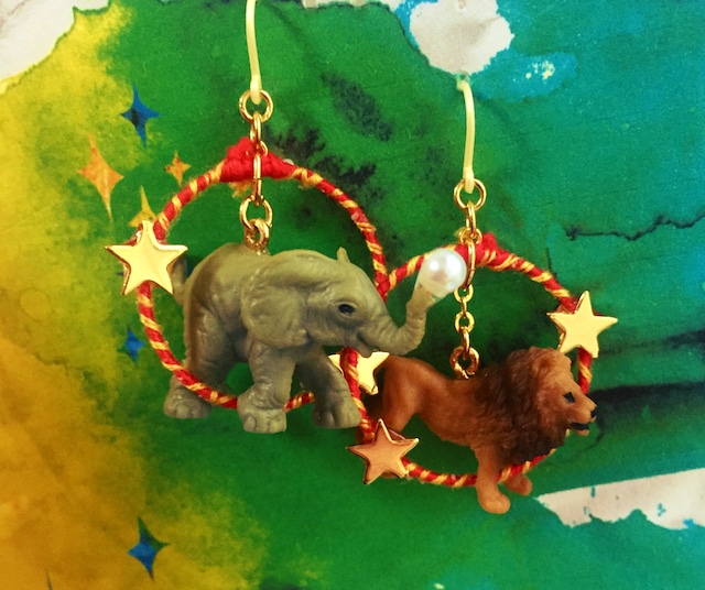 赤と黄金の糸で作ったリングを象とライオンが輪くぐりをしているようなデザインのピアスを写した写真。
