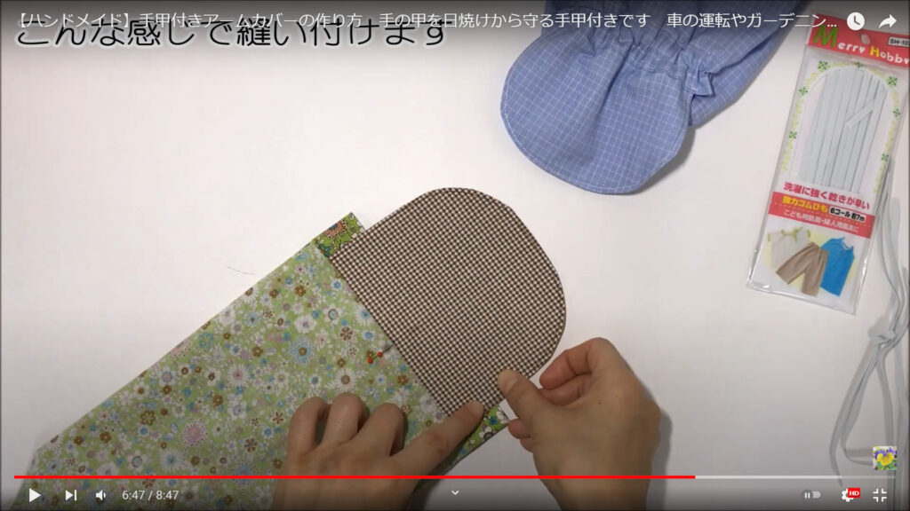 アームカバーの本体と手甲部分を縫い付ける作業を解説している動画で、「こんな感じで縫い付けます」という文と共に、作業台に置かれた本体と手甲部分を合わせた部分を手で指さしている画像。