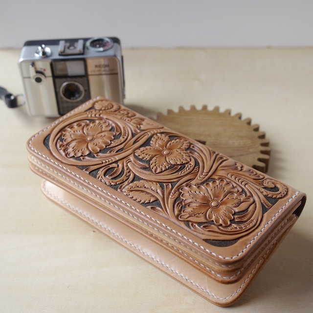 レザーカービングという細工の施された、革製の長財布の画像。財布は木製の歯車の上にディスプレイされており、表面には曲線を多く含んだ繊細な花の模様が浮かび上がっている。