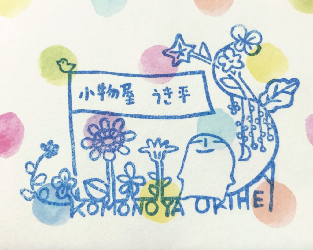 作家さんが運営されているネットショップのロゴ画像。淡い色の水玉模様を背景にして、花や店のキャラクターをあしらったデザインのスタンプが青色のインクで押されている。