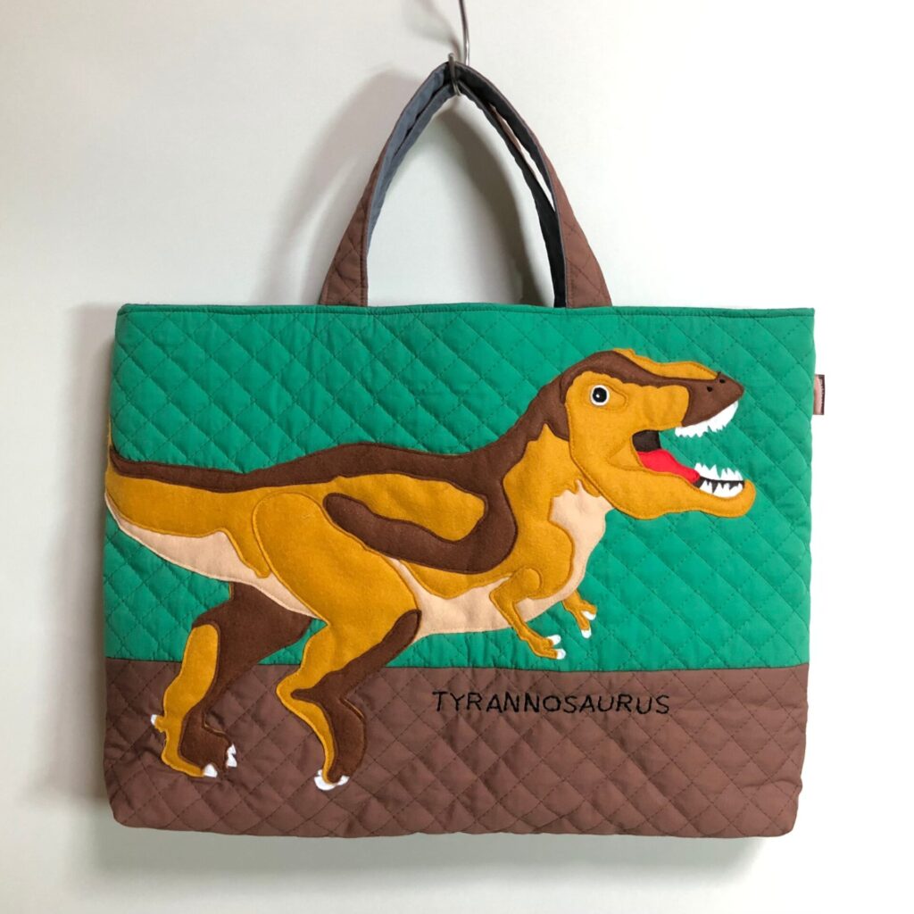 ティラノサウルスの手提げバッグ。下が茶色で上がエメラルドグリーンの生地。真ん中に黄色と茶色のティラノサウルス。