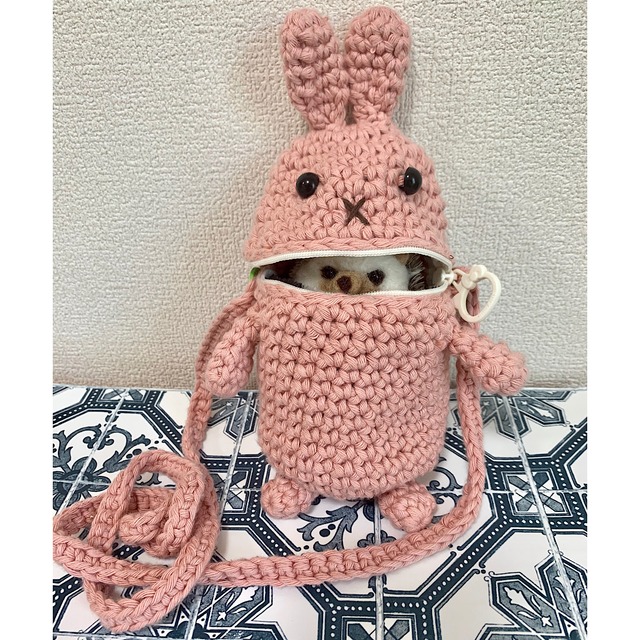 毛糸で編んだピンクのウサギのポーチ。中から白い顔の人形がのそいている様子。
