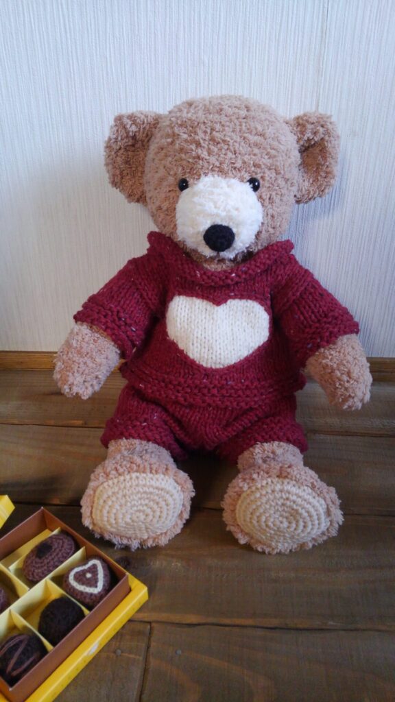 クマのぬいぐるみが座っている様子。真ん中にハートのある赤いセーターを着ている。
