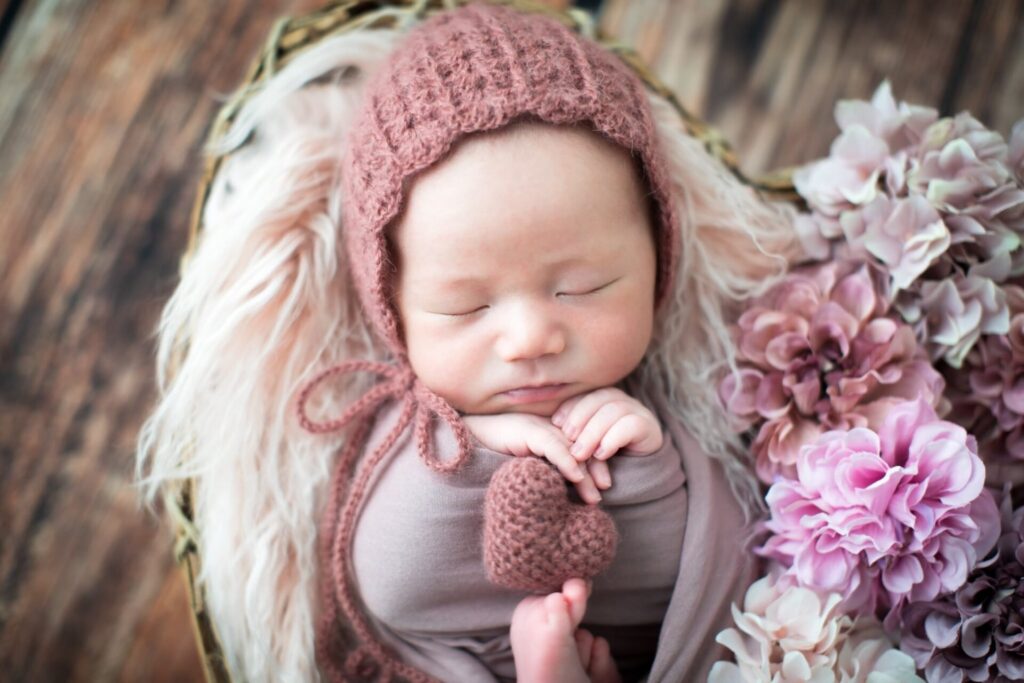 帽子をかぶった新生児がお花に囲まれて眠っている様子。
