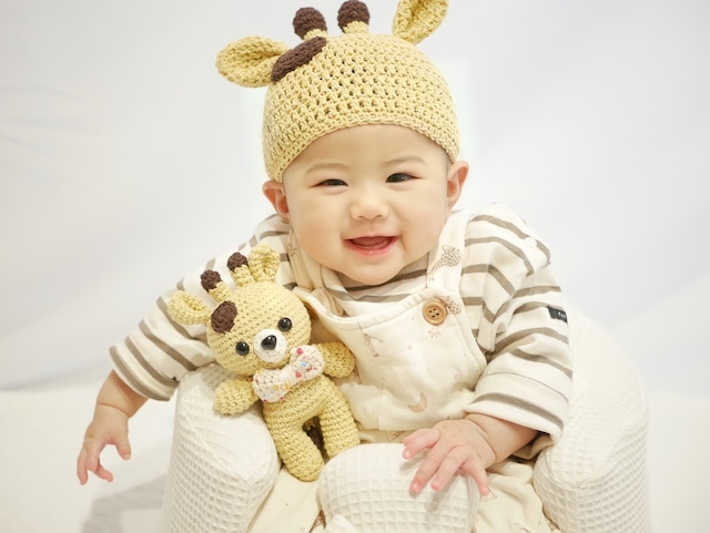 赤ちゃんがきりんのぬいぐるみを持ちながらキリンの帽子をかぶって笑っている様子。