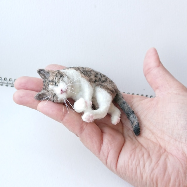 羊毛フェルトで作った猫が手の上で寝転がっている様子の写真。