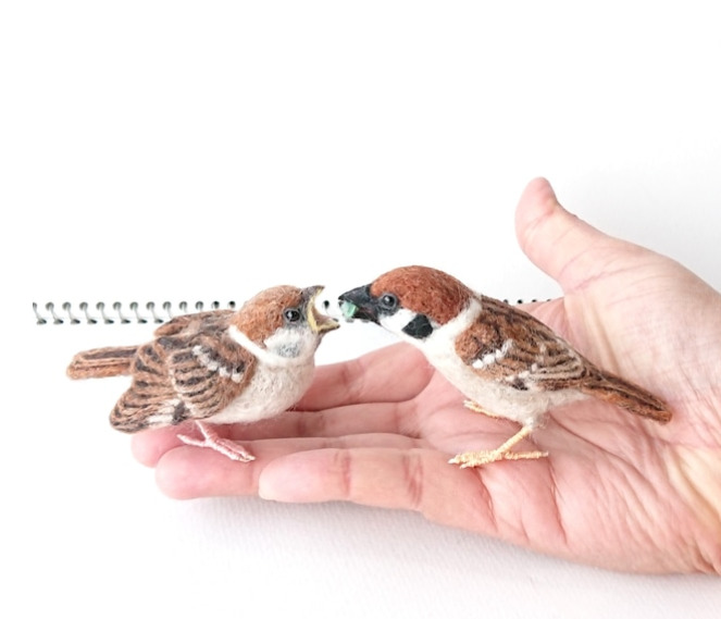 羊毛フェルトで作った二匹の雀が手のひらの上で餌を分け与えている様子の写真。