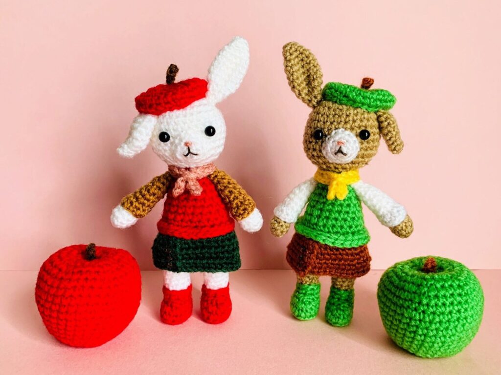 赤い服と赤いリンゴベレー帽、緑の服と緑のリンゴベレー帽をそれぞれ身に着けたウサギのあみぐるみが2点、並んで立っている画像。それぞれには赤と緑のリンゴのあみぐるみも添えられている。