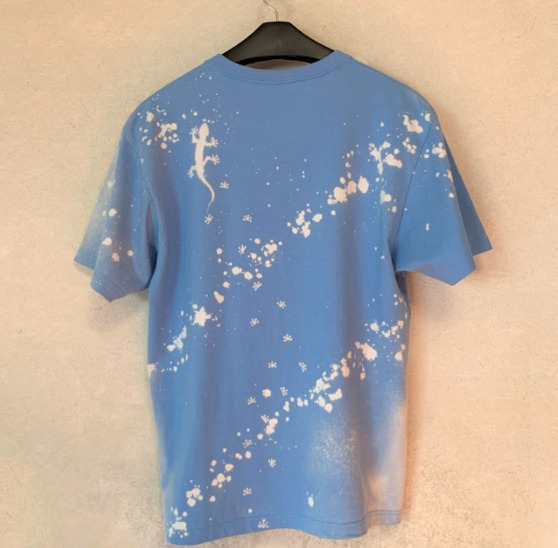 壁に、水色のTシャツがかかっている。Tシャツの背中左上にヤモリの絵が描かれている。Tシャツには斜めにブリーチ加工がされ、白抜きされている。