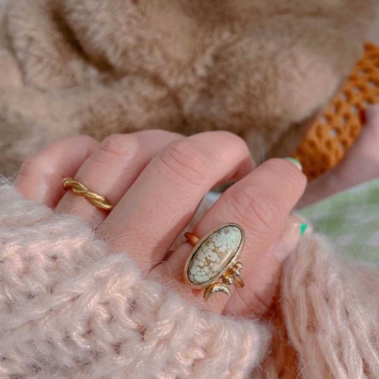 ニットを着た女性の手が写っており、人差し指にゴールド金具の指輪をしている。指輪には淡い白のターコイズがついている。