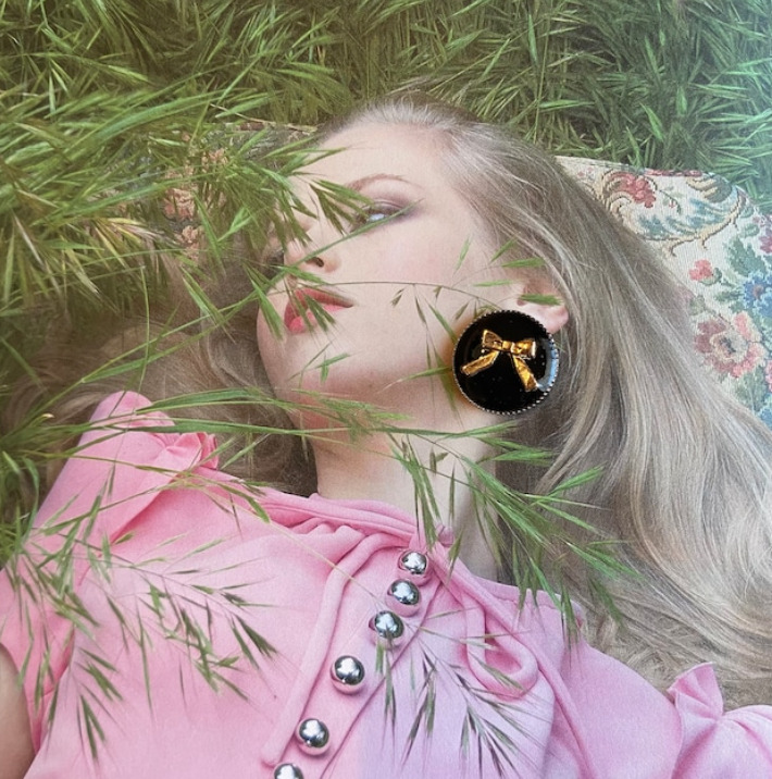 緑の中に寝そべる金髪の女性の写真の耳元に、ゴールドリボンが入った黒丸のイヤリングが置かれている