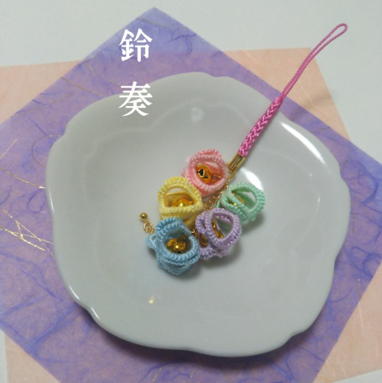 お皿の上にピンク、黄色、緑、紫、青の糸で編まれたストラップが置かれている。糸の編まれた中には鈴が入っている。