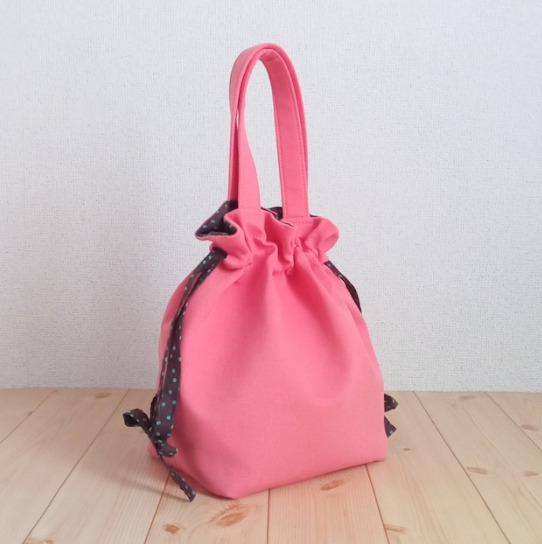 明るいピンクの巾着トートバッグが置かれている。紐はブラウンと水色のドット柄の生地が使われている。