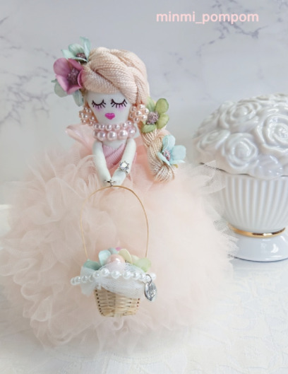 大理石柄の背景に、陶器のお花が飾られており、その手前にピンクのひらひらドレスを着た女の子の人形が飾られている。人形の黄色の髪にはお花が飾られており、パールのネックレスとピアスを着けている。カゴバッグを持っている。
