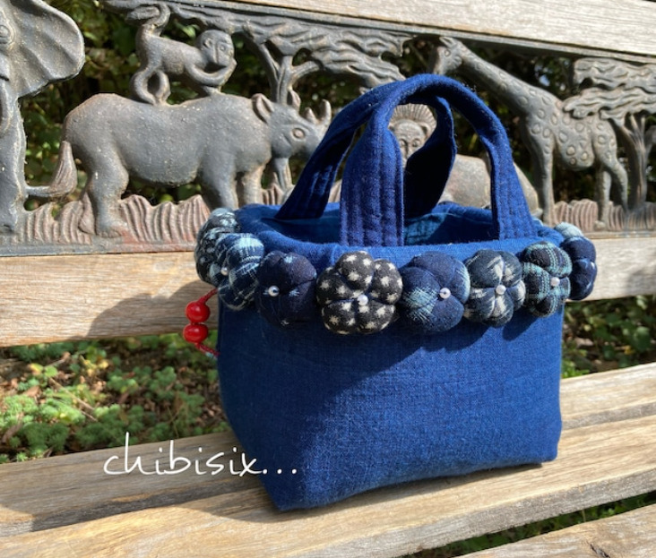ベンチの上にブルーの古布を使ったカゴバッグが乗っている。カゴバッグの周りにはお花のモチーフがついている。
