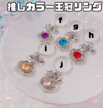 白い皿の上に、シルバーの王冠パーツのついたリングが五個置かれている。王冠の真ん中に紫、ブルー、赤、オレンジ、ピンクのビジューがついている。