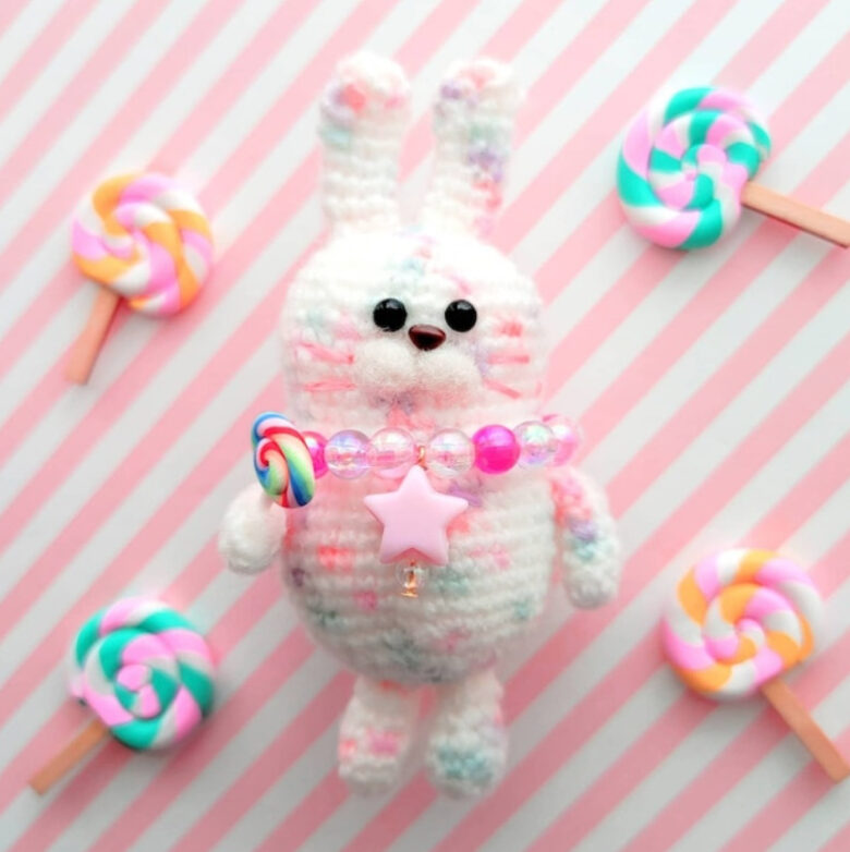 ピンクストライプの上にくるくるキャンディーとウサギの編みぐるみが乗っている。ウサギは白メインに、ピンクや紫、緑のカラーが混じっている。ピンクや透明、星パーツのネックレスを着けており、手元にはキャンディが握られている。