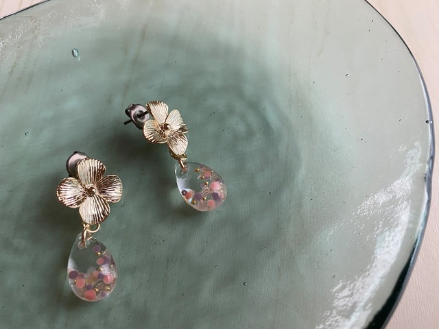 4弁の小さなゴールドカラーのお花から、雫がぶら下がるピアスの作品画像。透明なレジンの雫の中にはピンクやパープル、ゴールドのパーツが封入されている。