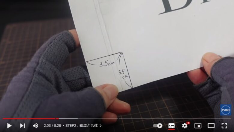 ディオールの紙袋のディオールと書かれた面をカットしたものを両手で持ち、その左隅に、縦3.5センチ、横3.5センチの黒い線が引かれている。