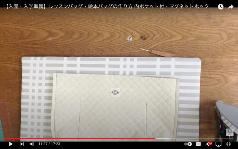 作業台の上に白とグレーの模様のアイロン代が置かれている。その上には作りかけのバッグが載っている。作業台の上部に、マグネットボタンとニッパーが置いてある。画面右下にはアイロンの一部が映っている。