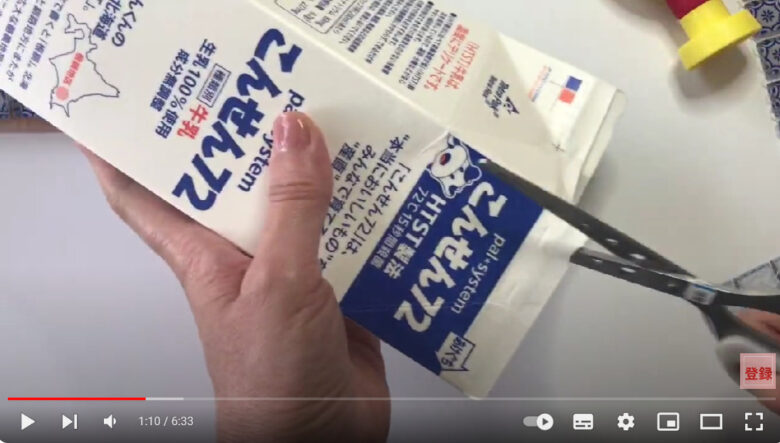 ガラスタイルのコースターで使用する牛乳パックのへらの作り方を説明している動画で、牛乳パックを手に持ち、ハサミでカットする様子を表示した画像。