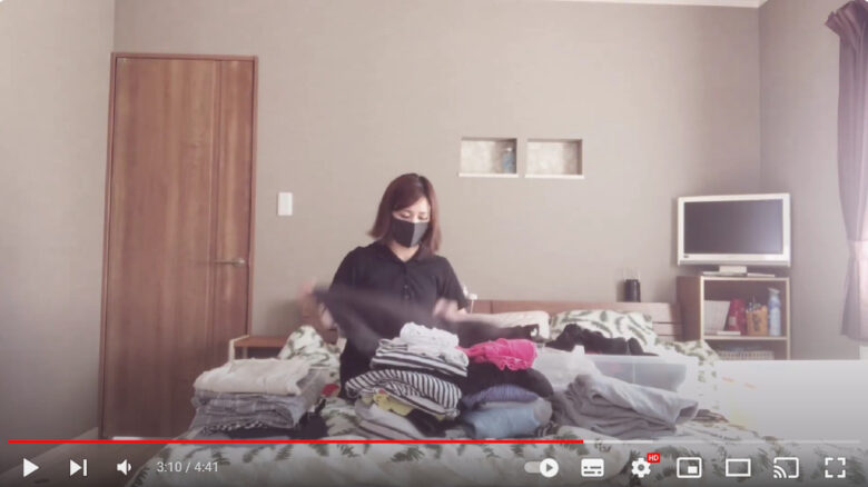 女性がベッドの上で多くの服を畳んでいる様子。女性の前には畳まれた服が山積みになっている。